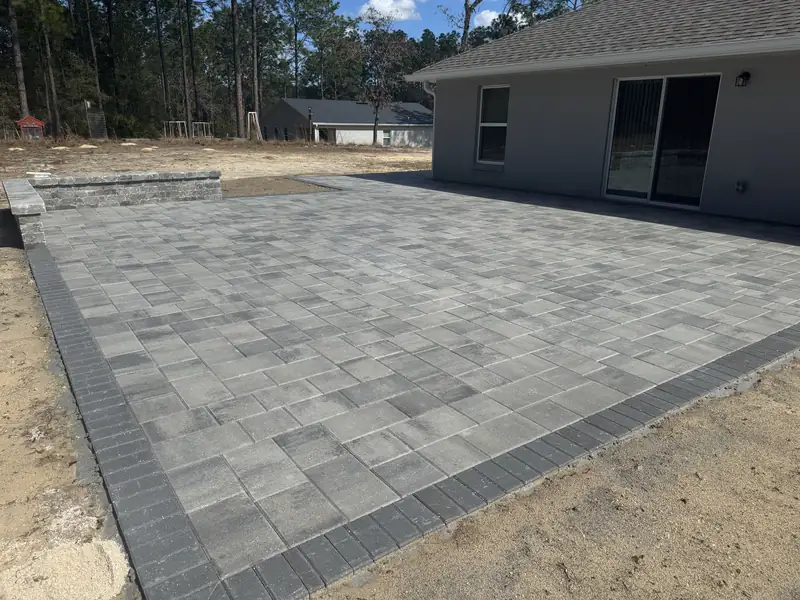 Large grey paver patio with a dark border and a low seat wall behind a new home in Dunnellon