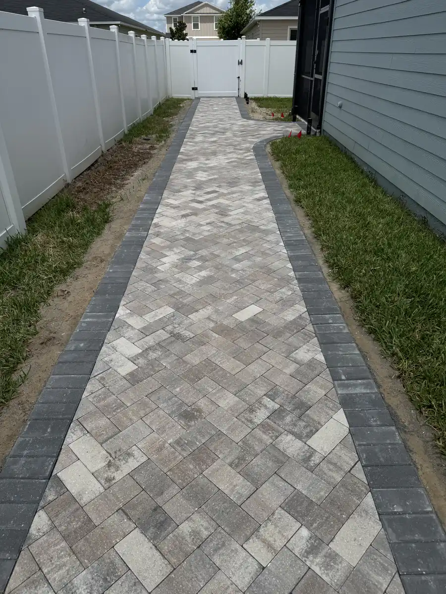 Long herringbone paver walkway with dark borders running beside a white vinyl fence to a side gate in Ocala
